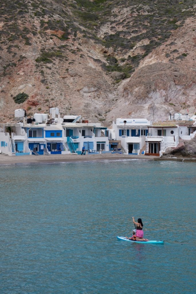 Paddleboarder crossing Firopotamos cove below waterfront boathouses