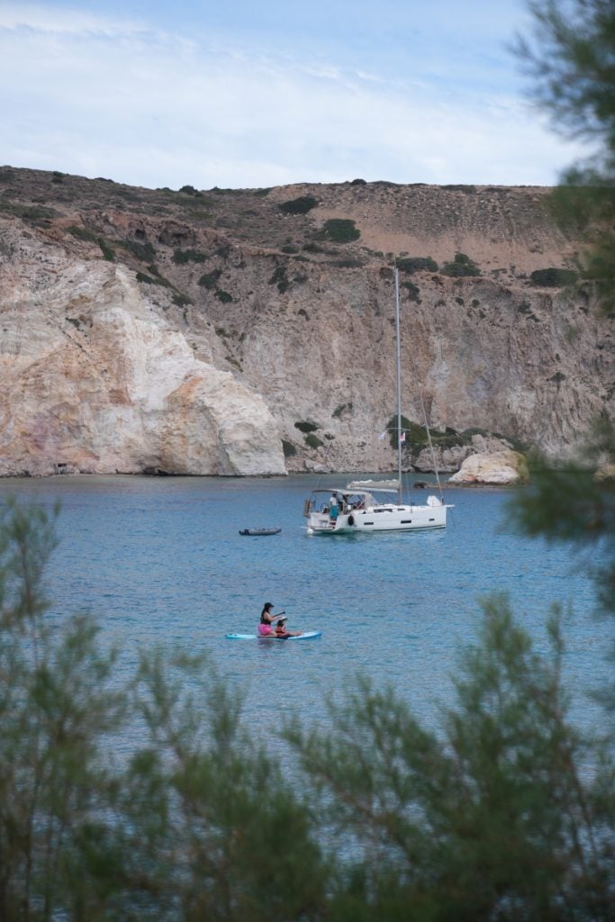 Paddleboarder passes a sailboat in a rocky Firopotamos cove
