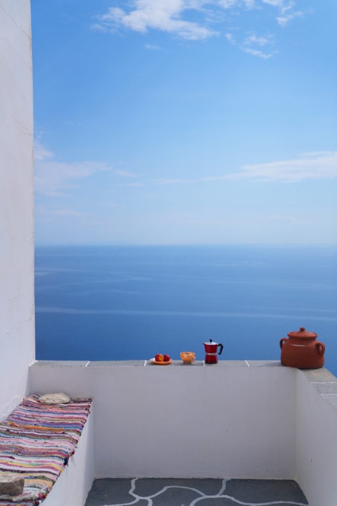 Sea view from a whitewashed terrace with a striped cushion and a red coffee pot.