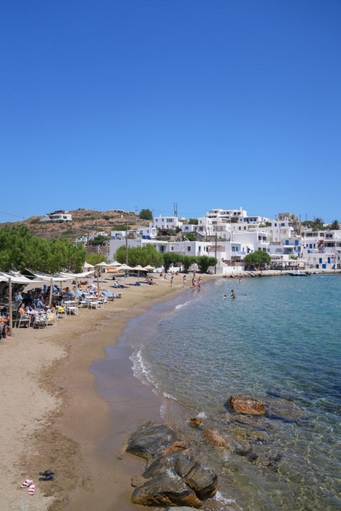 Faros beach with swimmers, waterfront buildings and shaded tables along the sand.
