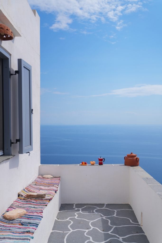 Whitewashed terrace with blue-gray shutters, a striped bench cushion and a sea view.